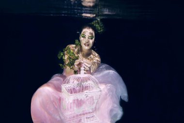 young beautiful woman in long  pink dress with cage in her hands underwater
