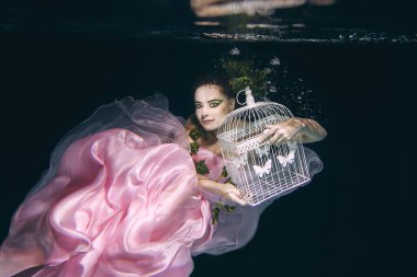 young beautiful woman in long  pink dress with cage in her hands underwater
