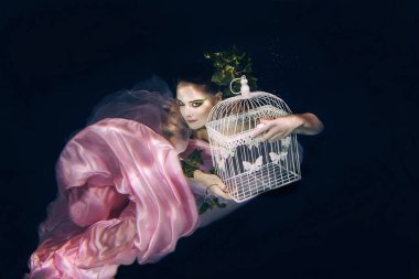 young beautiful woman in long  pink dress with cage in her hands underwater