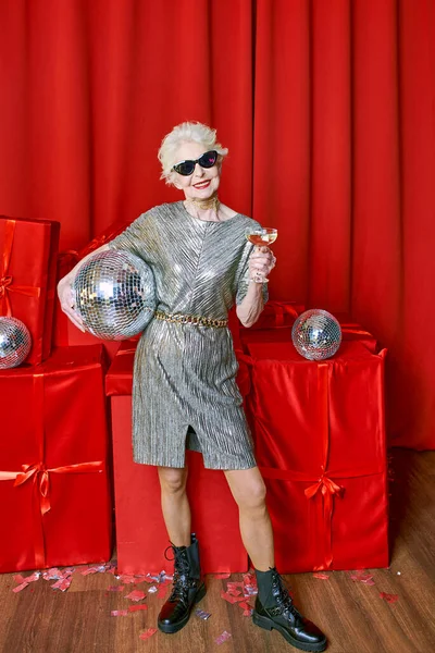 Senior stylish elegant woman with glass of sparkling wine on red background. Party, fashion, celebration, anti age concept