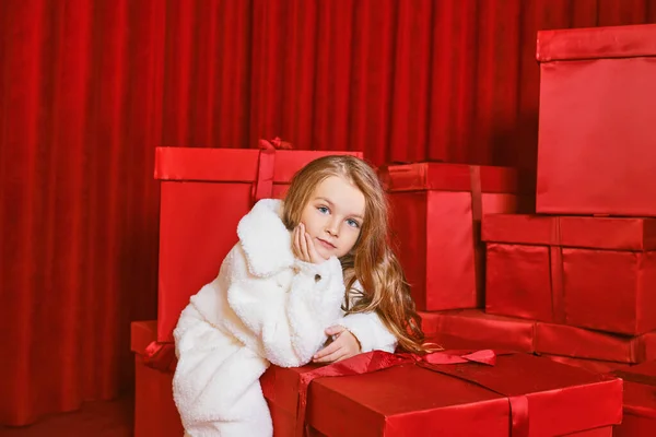 Cute little girl in white coat by the huge red christmas presents boxes on red background. Holidays, shopping, fun, fashion