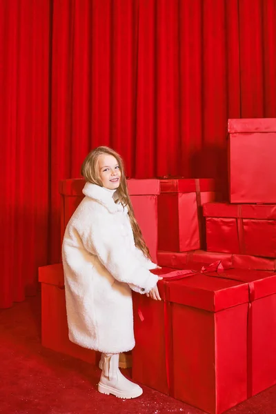 Cute little girl in white coat by the huge red christmas presents boxes on red background. Holidays, shopping, fun, fashion