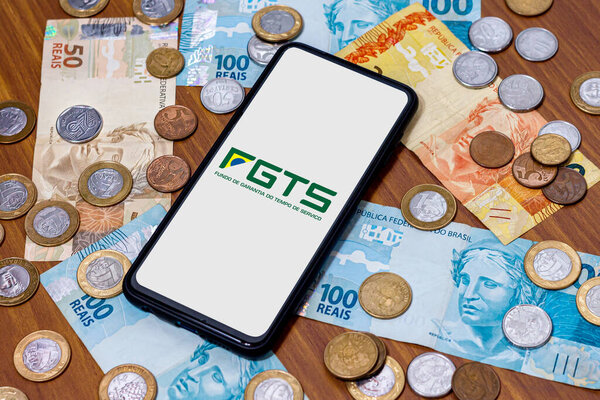 Sao Paulo, Brazil. SEPTEMBER 19, 2022: FGTS logo on smartphone screen with multiple coins and moneys around. FGTS is the Brazilian worker's guarantee fund. Payment by the Brazilian government