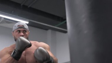 Sakallı güçlü genç erkek boks eldivenleriyle kum torbası tekmeliyor. Spor eğitimi sırasında yavaş çekimde eller ve bacaklar kullanıyor.
