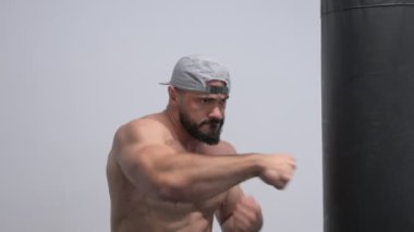 Spor salonunda boks antrenmanında kum torbasının yanında şapkalı güçlü, genç, sakallı, kaslı bir erkek.