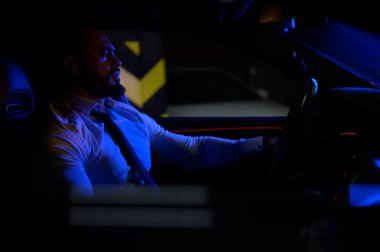 cute bearded strong young man in white shirt with tie sitting in luxury sport muscle car lit with blue light in underground garage at night city