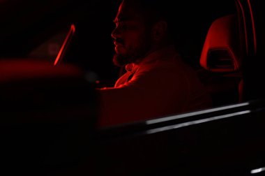 cute bearded strong rich young man in white shirt sitting in luxury sport muscle car lit with red light in night city