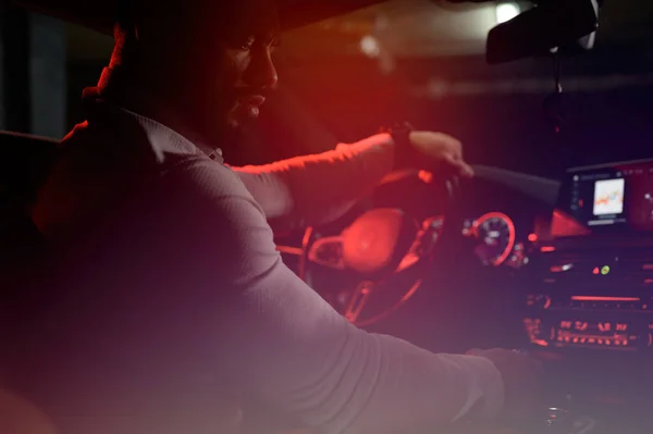 Cute bearded strong young man in white shirt sitting in luxury sport muscle car lit with red light in underground garage Fotografie de stoc