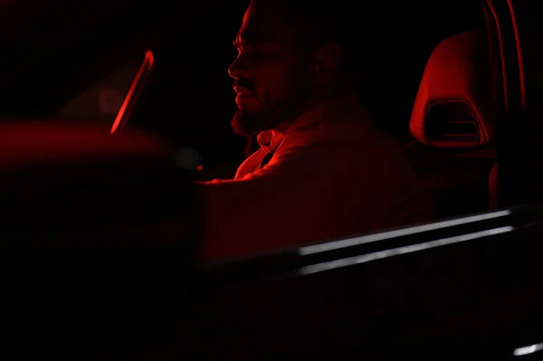 Cute bearded strong rich young man in white shirt sitting in luxury sport muscle car lit with red light in night city Immagine Stock