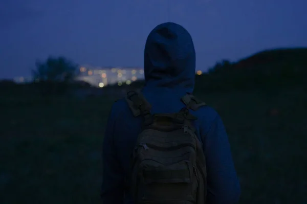 Om fără adăpost în hanorac cu rucsac uitam pe clădirile orașului de noapte în timpul asfințitului drumeții singur Fotografie de stoc