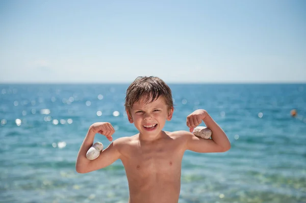 Šťastný rozkošný malý kluk ukazuje svaly s kameny na biceps na mořském pozadí Royalty Free Stock Fotografie