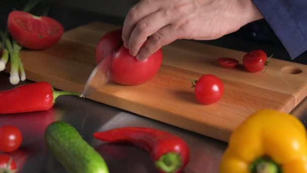 Primer plano de cocinero manos de cocina con cuchillo cortado tomate rojo en tablero de madera con muchas verduras alrededor — Vídeo de stock