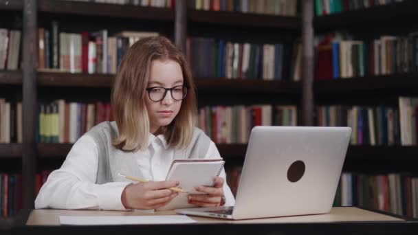 Καυκάσιος νεαρό κορίτσι φορώντας γυαλιά κάνει σχολική εργασία μελετώντας σε απευθείας σύνδεση χρησιμοποιώντας φορητό υπολογιστή κρατώντας σημειώσεις στο βιβλίο αντιγράφων, ενώ έχοντας μια βιντεοκλήση με θηλυκό δάσκαλο στο φορητό υπολογιστή στη βιβλιοθήκη — Αρχείο Βίντεο