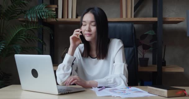 Conversación telefónica de negocios de mujer asiática gerente con un subordinado. Discusión de una nueva estrategia de negocio contra la oficina estilo loft telón de fondo. Mujer joven mira a la computadora portátil de pantalla — Vídeo de stock