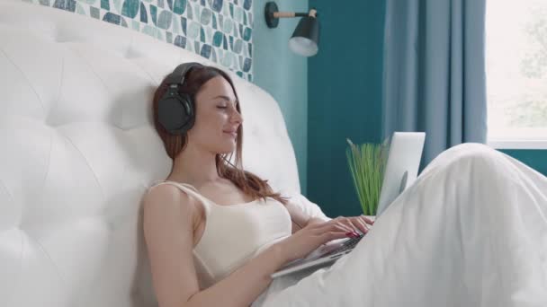 Selbstbewusste brünette Frau mit Kopfhörer tippt Nachricht in Computer auf dem Bett. Glückliche hübsche Frau arbeitet von zu Hause auf Laptop-Schlüsselcomputer aus dem Schlafzimmer. — Stockvideo