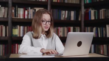 Genç kız öğrenci kolej öğrenimi internette öğretmenle el sallarken webcam 'in bilgisayardaki video konferans görüşmesini kullanıyor. Kütüphane arkaplanı üzerinden video konferansı eğitimi