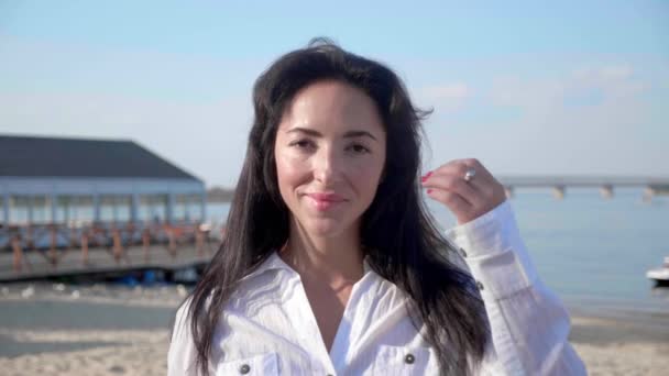 Portret młodej kobiety patrzącej w kamerę stojącej na plaży z morzem na tle. Atrakcyjna dziewczyna w białej koszulce podnosi oczy. Wiatr wieje przewiewne brązowe włosy świecące w słońcu flary. — Wideo stockowe