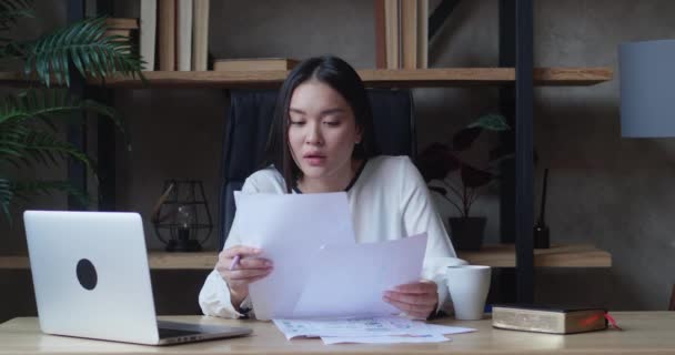 Азиатская деловая женщина от усталости закрывает ноутбук ставит документы в сторону и опирается на спинку кресла — стоковое видео