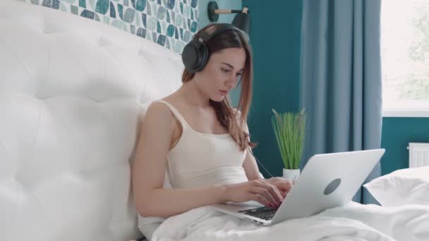 Femme brune dans le message de dactylographie casque dans l'ordinateur sur le lit. Jolie jolie femme travaillant de la maison sur ordinateur portable clé de la chambre. — Video