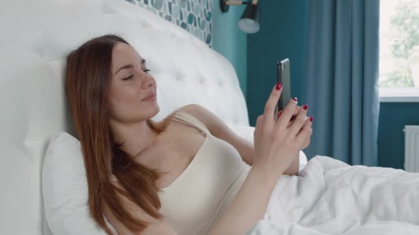 Ontspannen jonge brunette vrouw liggend op wit bed met smartphone in handen. Aantrekkelijk meisje ontspannen thuis met moderne gadget. — Stockvideo