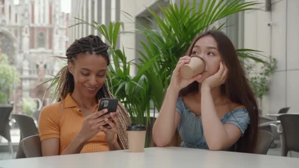 スマホを持っている混合レースの若い女の子たちは、屋外のカフェに座って指を指している間、コーヒーショップでソーシャルメディアアジアの女性はコーヒーを飲みます。インターネットの概念 — ストック動画