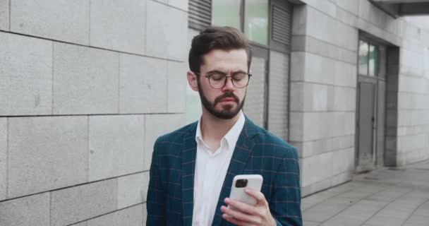 Důvěryhodný mladý podnikatel v brýlích procházení mobilní telefon internet během přestávky chůze podél obchodní čtvrti. Úspěšný podnikatel jde do práce. — Stock video