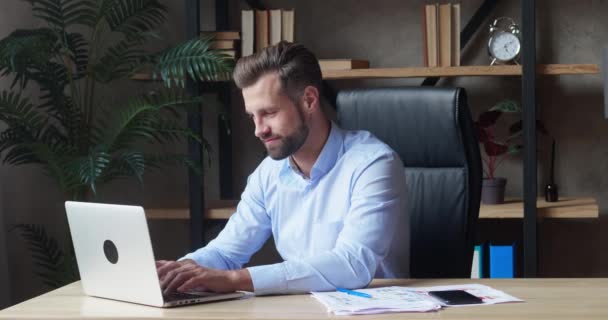 Giovane uomo d'affari felice in camicia blu guardando nel computer portatile, digitando testo, godendo pausa di lavoro sorridente seduto alla scrivania magra sul retro di una sedia. Sentire un senso di soddisfazione professionale — Video Stock