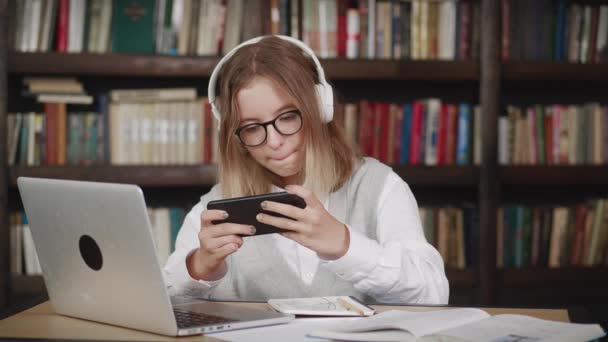 幸せな女性の10代のゲーマーは、ライブラリ内のスマートフォンとオンラインヘッドフォン競争プレイビデオゲームを着用してください。オンラインスポーツストリーミングゲーム。勝利の概念 — ストック動画