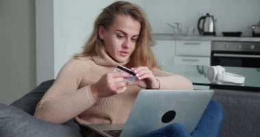 Sarışın kadın elinde banka kartı tutarak online ödeme yapıyor evdeki modern dizüstü bilgisayarla kanepede oturuyor ve gülümsüyor. Finans, internet ve gençlik konsepti alışverişi. Çevrimiçi kredi kartı kullanılıyor