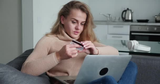 Блондинка делает онлайн-платеж, держа банковскую карту, используя современный ноутбук дома, сидя на диване и улыбаясь. Финансы, покупки в Интернете и молодежной концепции. Использование кредитных карт онлайн, онлайн — стоковое видео