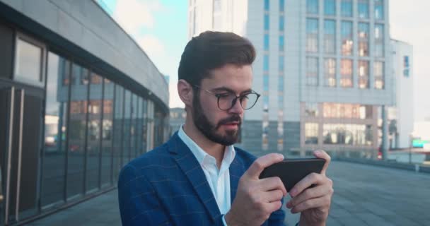 ビジネスマンはスマートフォンでビデオゲームをしている。都会の背景に対してオンラインゲームをプレイハッピーオフィスの男。勝利の概念. — ストック動画