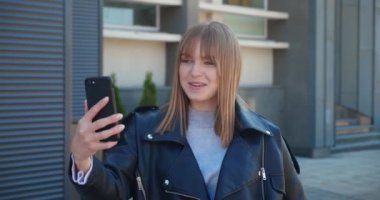 Neşeli, çekici sarışın kadın akıllı telefondan konuşuyor ve şehirde kameraya el sallıyor..