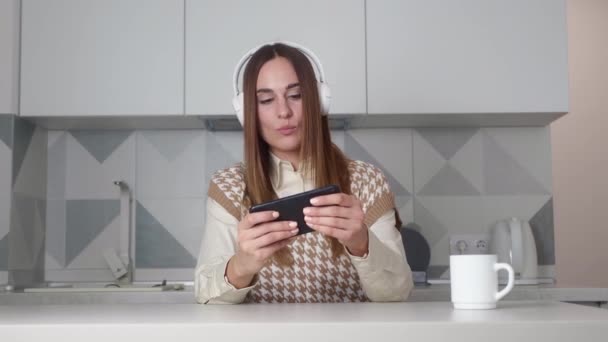A pleased young woman is playing games using his smartphone while holding it horizontally standing sitting over the kitchen background. — стоковое видео