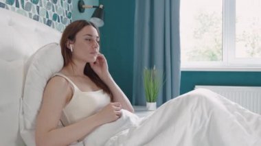 Kendine güvenen genç bir kadın, mutlu bir kulaklık takıyor yatak odasında rahatlıyor ve müzik dinliyor, kadın eğlencesi yapıyor ve akıllı cep telefonuyla şarkı çalıyor, yaşam tarzı kavramı..