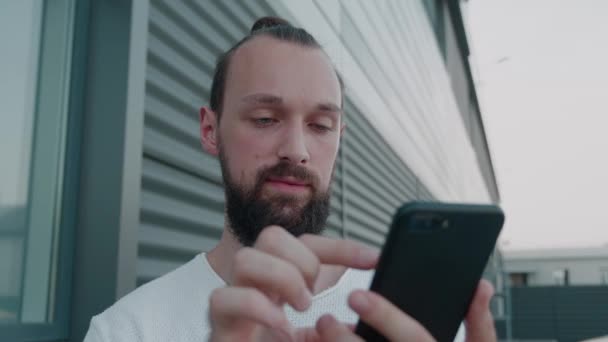 Молодий бородатий хіпстер дивиться на екран смартфона і вирішує купити онлайн або перевернути через стрічку новин — стокове відео