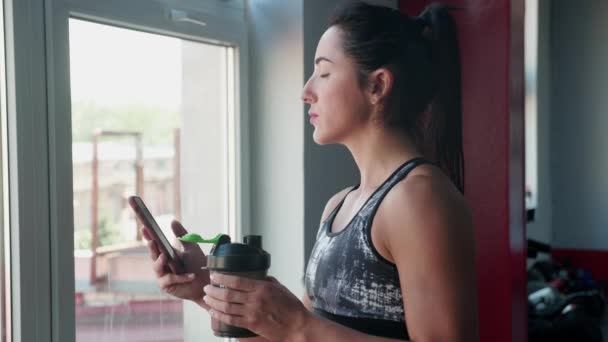 Mujer joven en traje deportivo está de pie cerca de la ventana mirando en el teléfono inteligente y bebe bebidas multivitamínicas por la mañana, cuidando de su salud, disfrutando del clima soleado en el gimnasio. — Vídeos de Stock