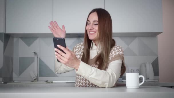 Онлайн-конференция из дома на кухне. Молодая красивая женщина европеоидного цвета в жилете с отпечатком говорит по видеосвязи с помощью мобильного телефона и эмоционально счастлива — стоковое видео