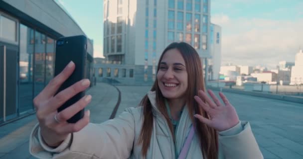 Веселая привлекательная рыжая женщина разговаривает на видео на смартфоне и машет перед камерой в городе. — стоковое видео