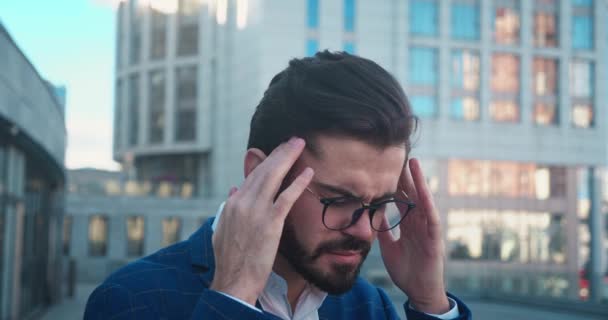 Stressed young man in glasses suffering from muscles tension, having painful head feelings due to. Tired employee overwhelmed with tasks near bussinescenter standing in outdoor. — стоковое видео