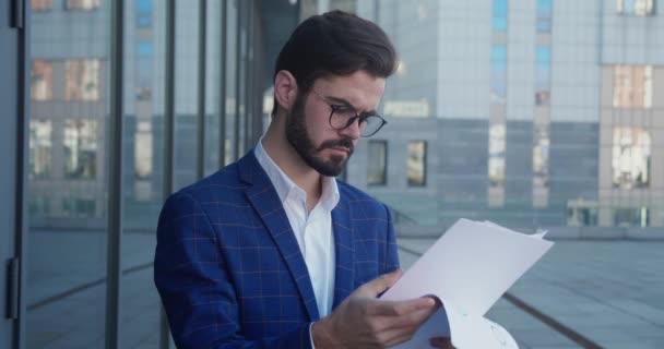 Young businessman in a formal suit and glasses with a serious face examines documents on the street. — Stockvideo
