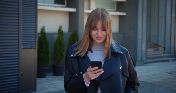 Senhora sorrindo verificando smartphone roupa urbana moderna, jaqueta de couro. A menina está olhando para a tela do telefone. — Vídeo de Stock