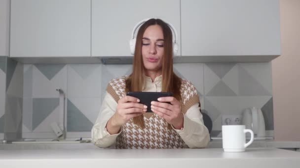 Жінка в навушниках грати з смартфоном на сучасному фоні кухні . — стокове відео