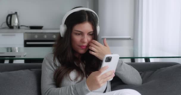 Atractiva chica adolescente morena en auriculares sentados del sofá en casa usando el teléfono y sonriendo. Me siento feliz. Comunicación mensaje de aspecto femenino en el teléfono celular o teléfono inteligente — Vídeos de Stock