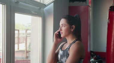 Spor kıyafetli genç bir kadın pencerenin yanında durup telefonla konuşuyor ve sabahları multivitamin içiyor. Sağlığıyla ilgileniyor, spor salonunda güneşli havanın keyfini çıkarıyor..