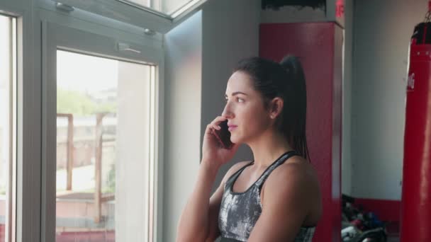 Young woman in sport outfit is standing near the window talking on the phone and drinks multivitamin beverage in the morning, taking care of his health, enjoying sunny weather in gym. — Stock videók