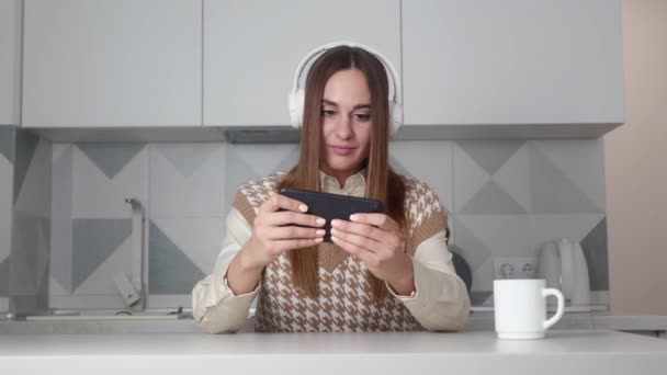 Amazed woman playing smartphone shocked, saying WOW. Female in headphone surprised to camera over kitchen background. — Stockvideo