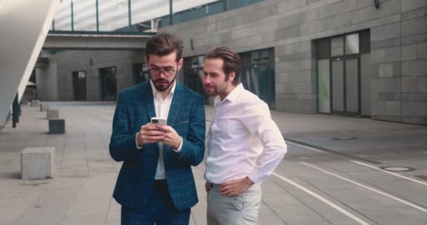 Caucasian young good looking man in business style tapping on his smartphone and showing something on it to his friend and business partner or colleague outdoor in the city center — Video Stock