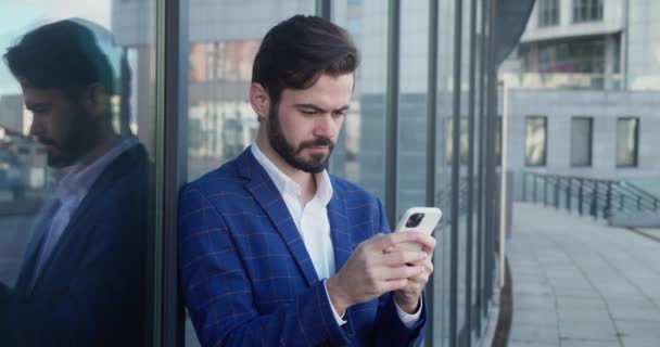 千年男性专业人员在玻璃大楼附近手持现代智能手机发短信. — 图库视频影像