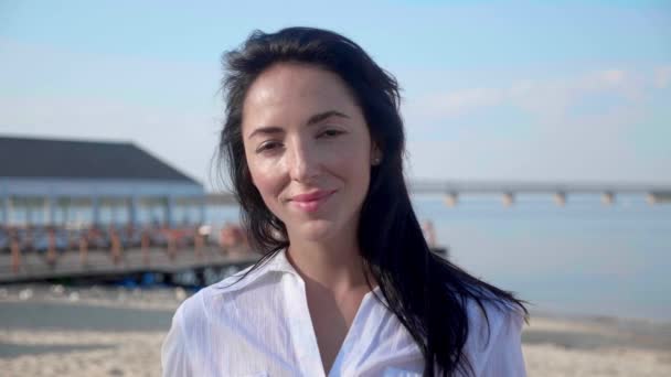 Portrait of young brunette woman looking at camera standing on the beach with a sea on background. Attractive girl in white t-shirt raises eyes. Wind blows airy brown hair shining in sun flares — Stockvideo