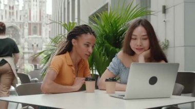 Asyalı kızlar ve Afrikalı Amerikalı öğrenciler üniversite kampüsünde dizüstü bilgisayarla tartışıyorlar - genç insanların öğrenme teknolojisini kullanması kavramı - genç bir kadının meslektaşına bakışıyla açıklaması